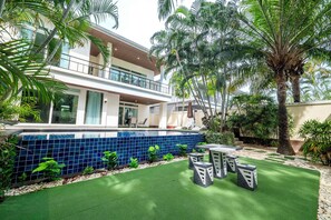 Ein modernes, zweistöckiges Gebäude mit Balkon, Pool mit blauen Fliesen und einer Terrassenanlage mit Kunstgrashalm und Sitzgelegenheiten.