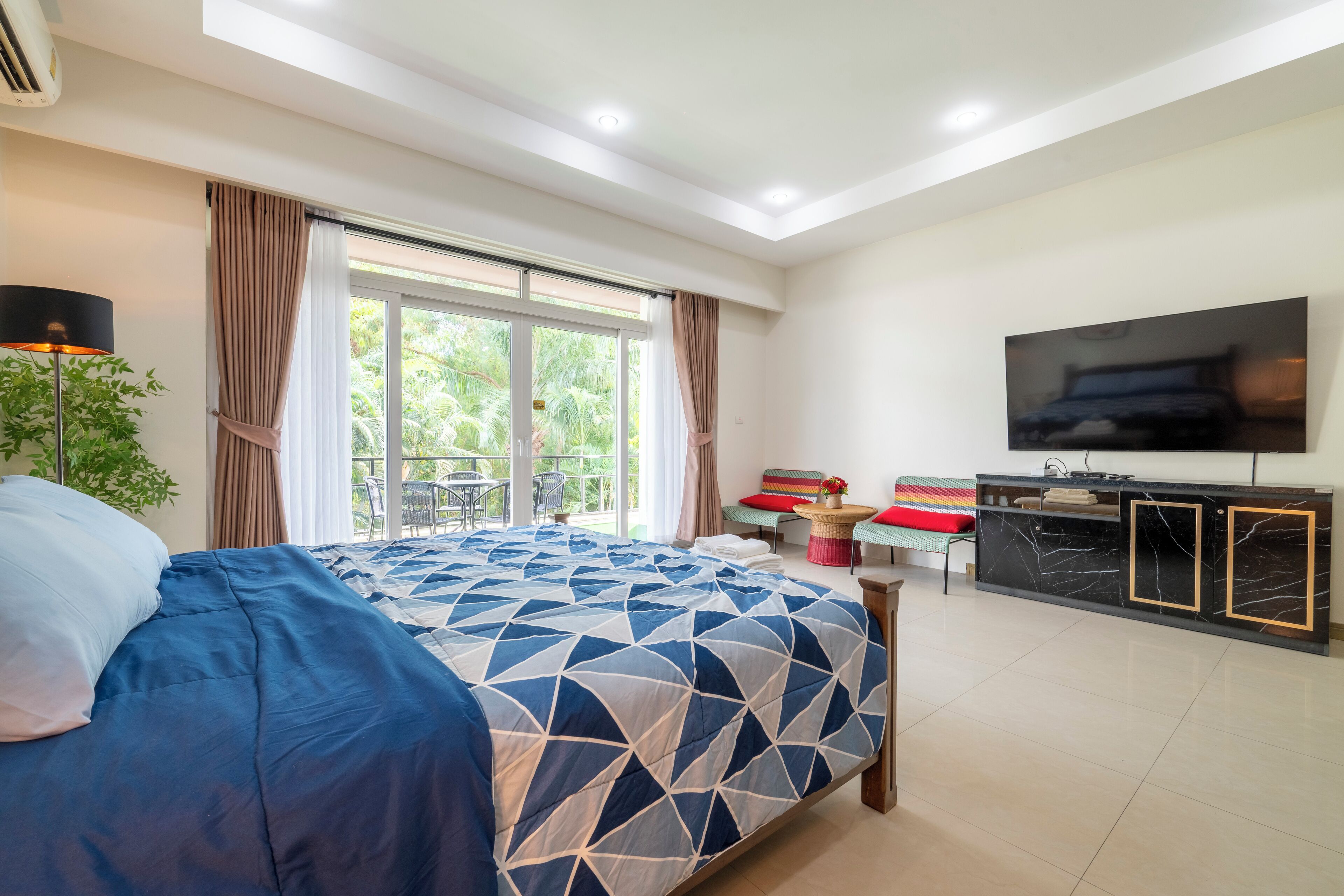 A modern bedroom with a large bed, a TV mounted on the wall, and a balcony with a view of greenery.