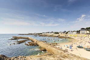 Uma praia costeira com um paredão rochoso, areal arenoso e vários barcos.