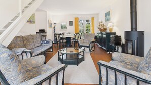 Un salon avec des canapés à motifs, une table basse et une cheminée. Un escalier se trouve à gauche, et un coin repas est visible au fond.