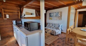 Cocina compacta con microondas, cafetera y refrigerador pequeño. Hay una mesa de comedor de madera con sillas y un sofá con funda estampada.