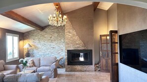 A living room with a stone wall, a fireplace, a chandelier, and a large TV. - Maison Belle Vue  – Charming villa with pool & breathtaking views - Near Nice (Carros)