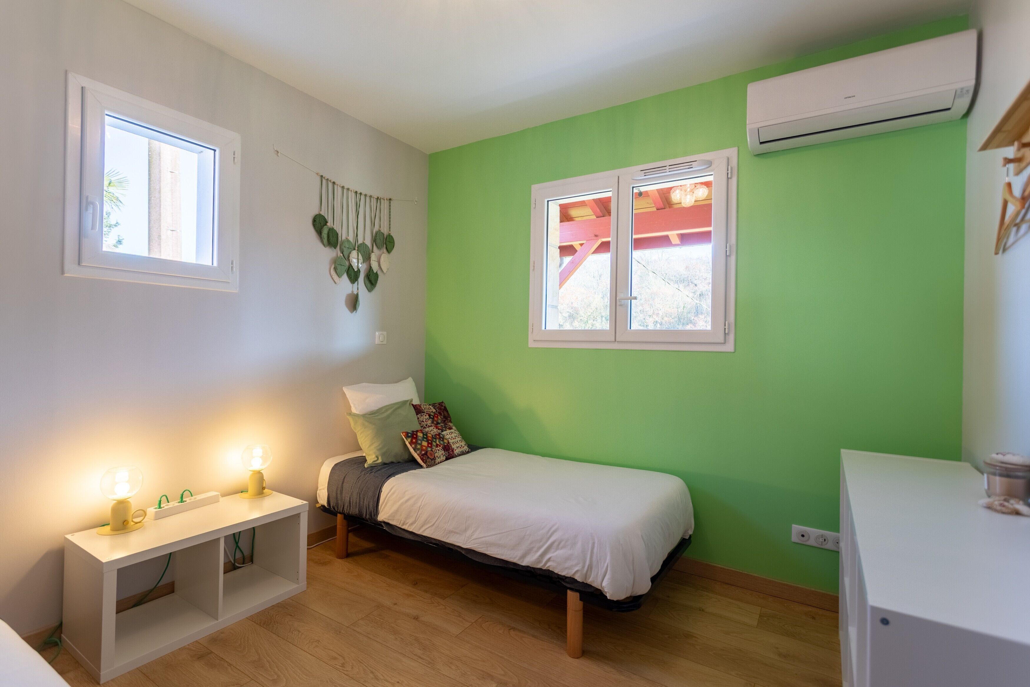Une chambre à coucher comprenant un lit, une table de chevet, deux fenêtres, un appareil de climatisation et une plante fixée au mur.