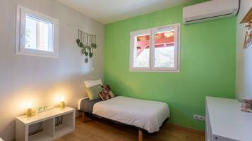 Une chambre à coucher comprenant un lit, une table de chevet, deux fenêtres, un appareil de climatisation et une plante fixée au mur.