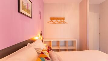 A bedroom with a bed, a bedside table, a hanging light, a wall-mounted shelf, and a door.