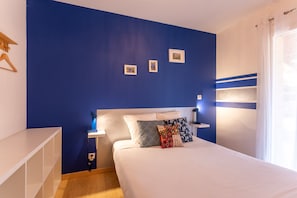 Une chambre avec un mur d’accent bleu, un lit blanc agrémenté d’oreillers à motifs, une table de chevet avec une lampe et une petite étagère avec des photos.