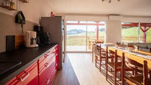 Une cuisine moderne dotée d’un grand îlot central, d’une machine à café et d’un réfrigérateur. Une porte-fenêtre coulissante donne accès à un balcon offrant une vue sur la campagne.