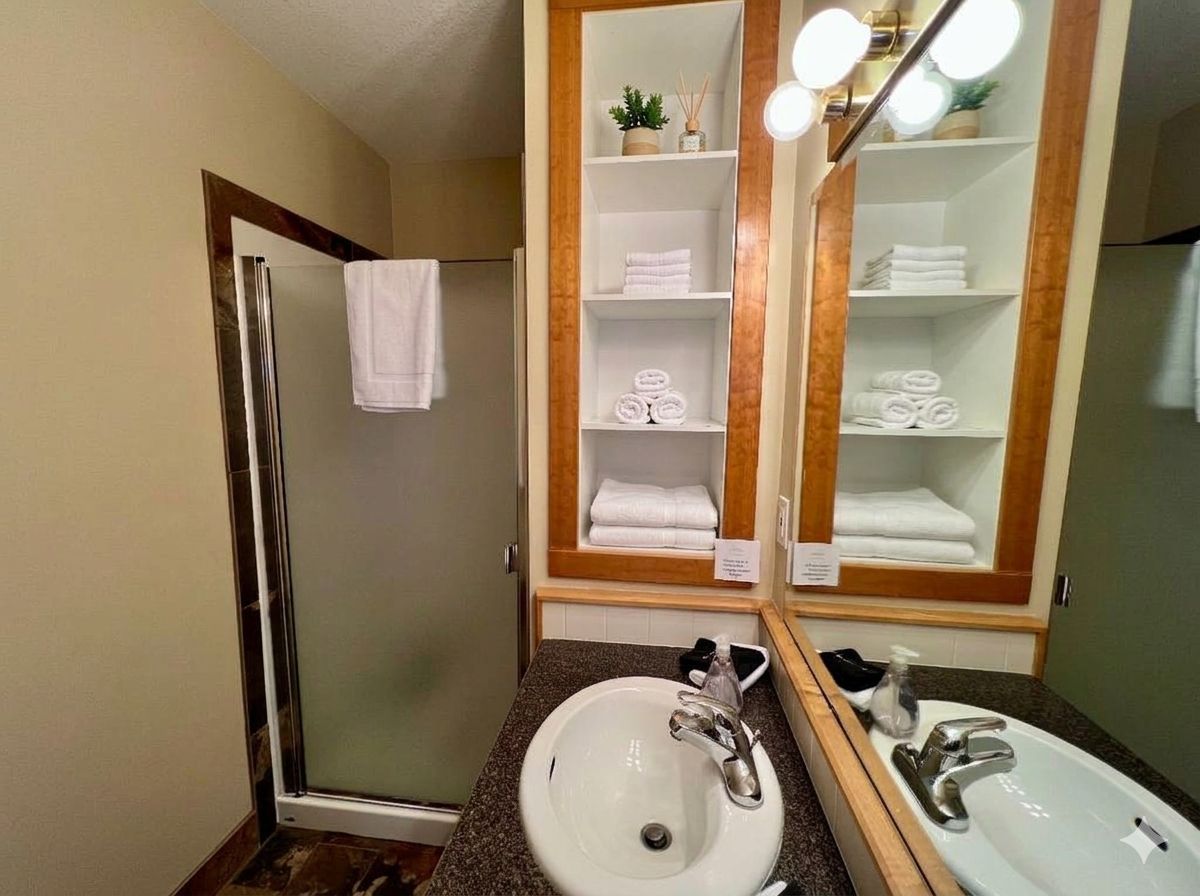 Un baño de hotel con cabina de ducha de vidrio, dos lavamanos y un espejo grande con repisas superiores.