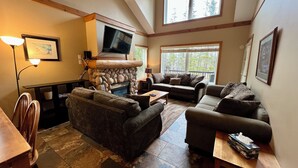 A spacious living room with a stone fireplace, comfortable sofas, a flat-screen TV, and large windows offering a view of trees. - Prime Base Location! 5BR, 4BA condo just a 2-min walk to the Express Quad. Ski back to your door. Features 2 hot tubs and a double garage for gear. Enjoy easy access to base restaurants and bars! (Kimberley)