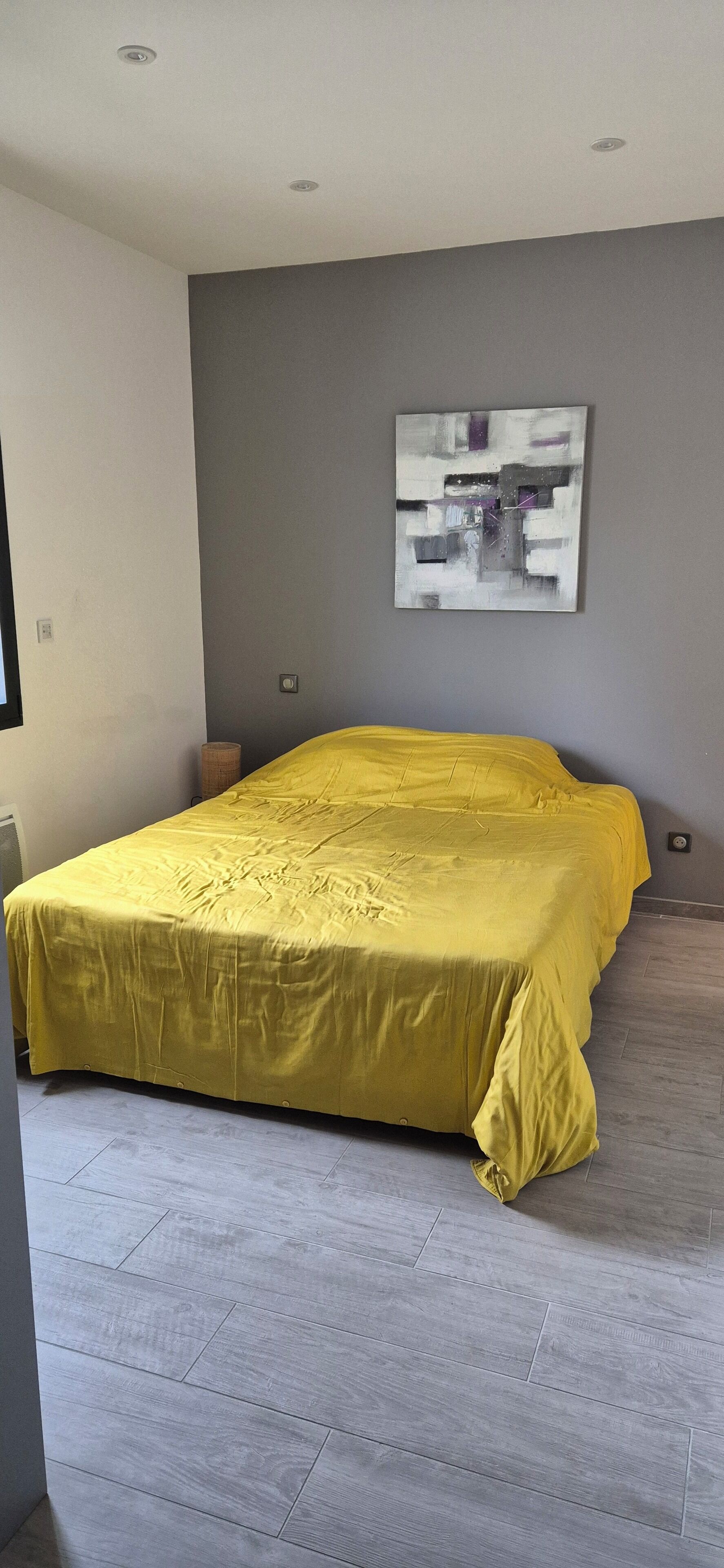 A bedroom with a bed covered in yellow bedding, a gray wall, and a modern abstract painting.