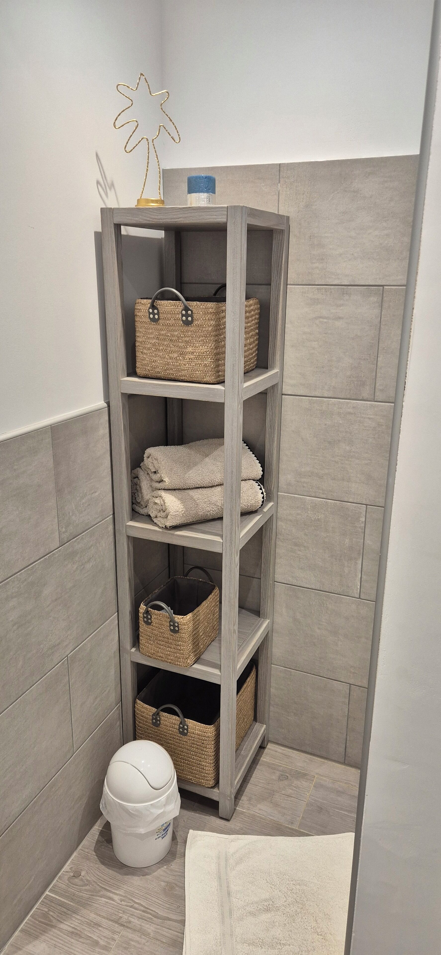 Un baño con pared de azulejos grises, un estante de madera con cestas tejidas y un cubo de basura blanco.