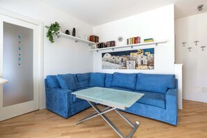 A room with a blue sofa, a glass-top table, a white door, and a wall-mounted shelf with books and a painting. - Appartamento Torretta 4 -Immobili e Soluzioni Rent (Bergamo)