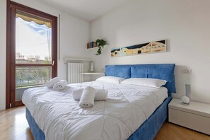 A bedroom with a large bed, a bedside table, a nightstand, a radiator, a window with a view, and a wall-mounted artwork. - Appartamento Torretta 4 -Immobili e Soluzioni Rent (Bergamo)