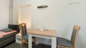 Een kleine kamer met een bed, een bureau met stoelen, een kapstok en een wandlamp.