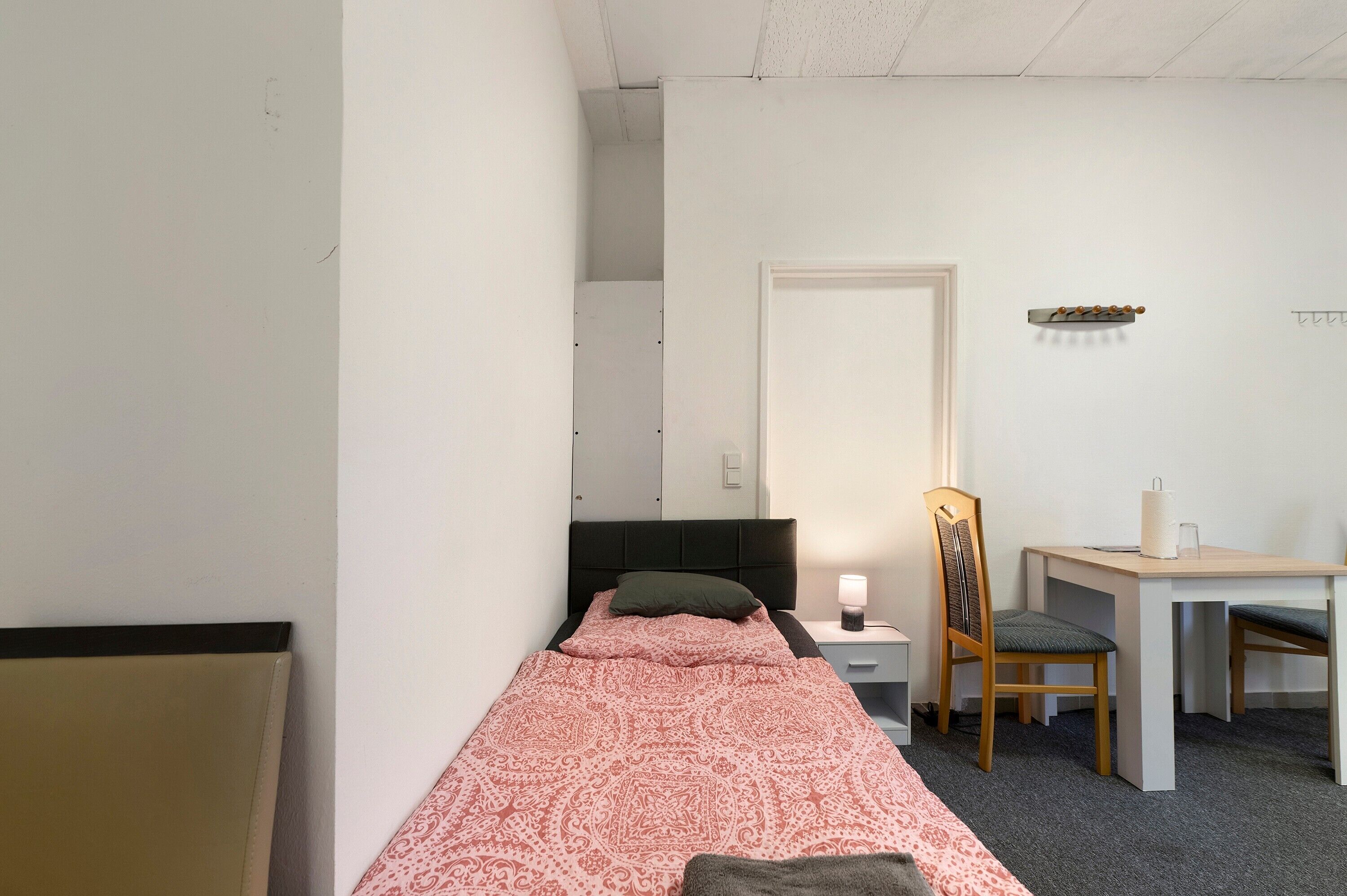 Ein kleines, schlichtes Zimmer mit einem Einzelbett, einem Schreibtisch, einem Stuhl und einem Nachttisch.