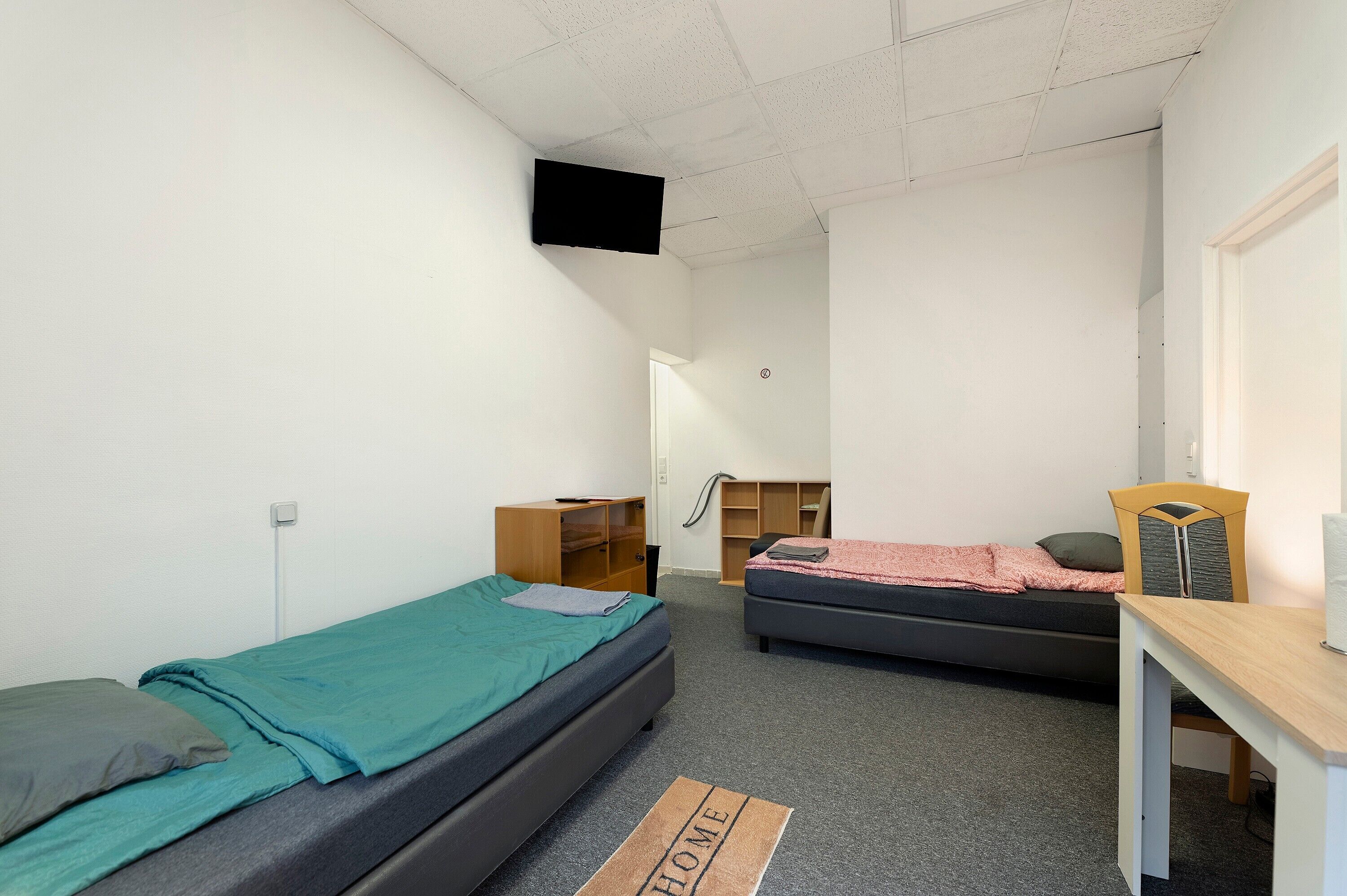 A dormitory room with two single beds, a desk, a chair, a TV mounted on the ceiling, and a wardrobe.