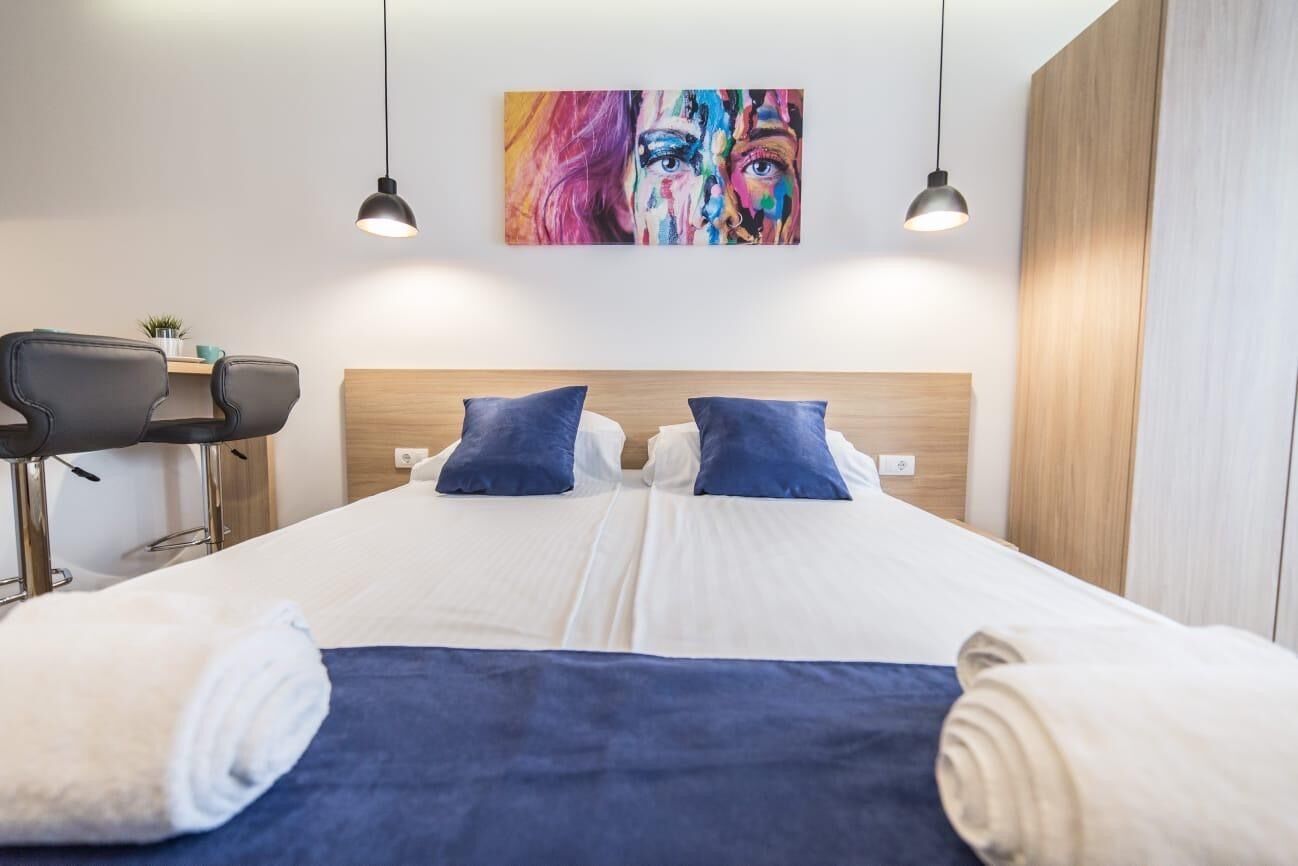 Una habitación de hotel moderna con una cama, dos almohadas, un escritorio con una silla, un cuadro en la pared y lámparas colgantes.