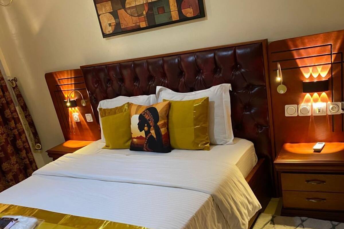 A hotel room with a large bed, a headboard with built-in lighting, a bedside table with a lamp, and a wall-mounted artwork.