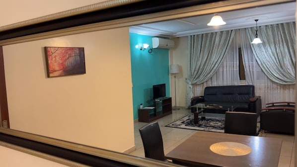 A living room with a teal accent wall, a black sofa, a coffee table, and a TV. There is a painting on the wall and a dining area with a table and chairs. - Appart'hôtel Micheline et Emile, Suites & SPA, Bastos (Yaoundé)