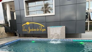 Una piscina con bordo piastrellato, un elemento decorativo d'acqua e un edificio con il cartello 'Les Jardins de Lym'.