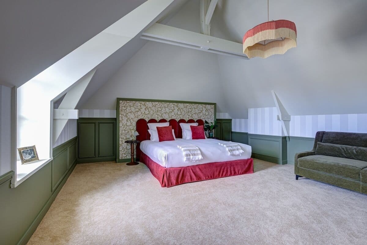 A bedroom with a large bed, a green sofa, a hanging light fixture, and a window with a picture frame.