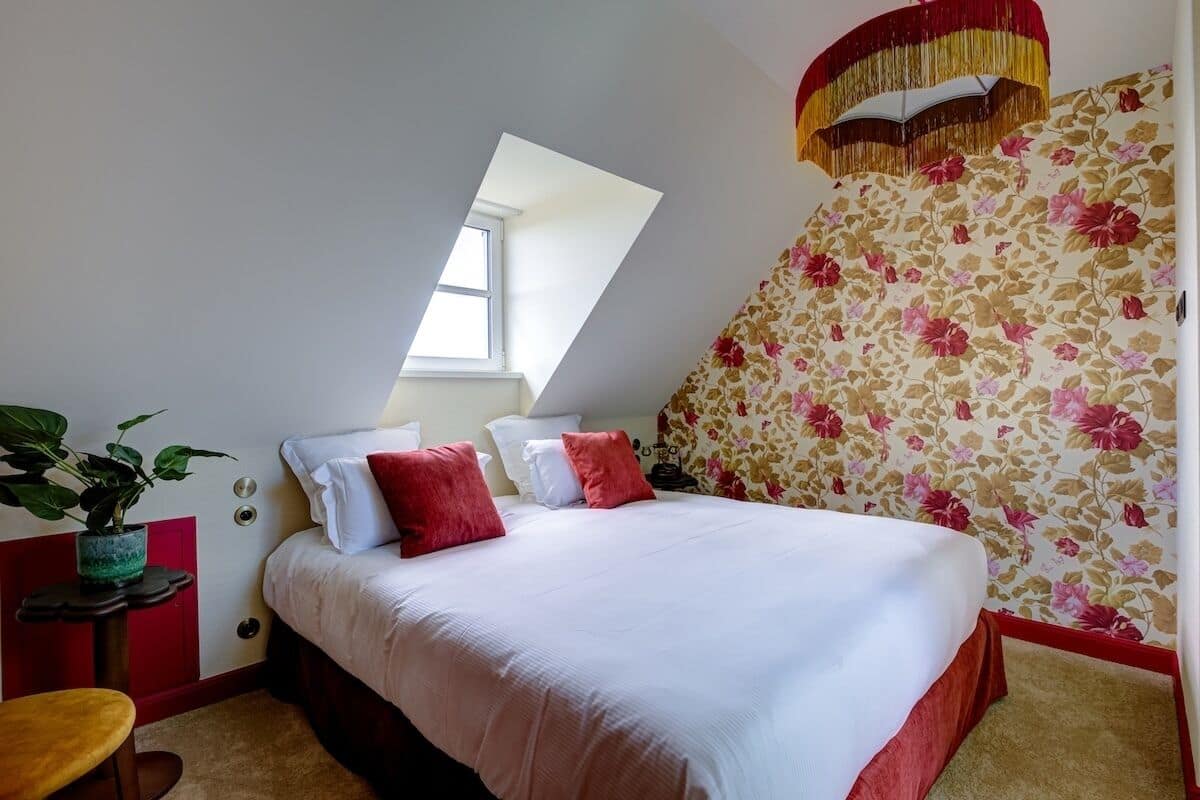 A bedroom with a bed, a window, a plant, and a wall with floral wallpaper.