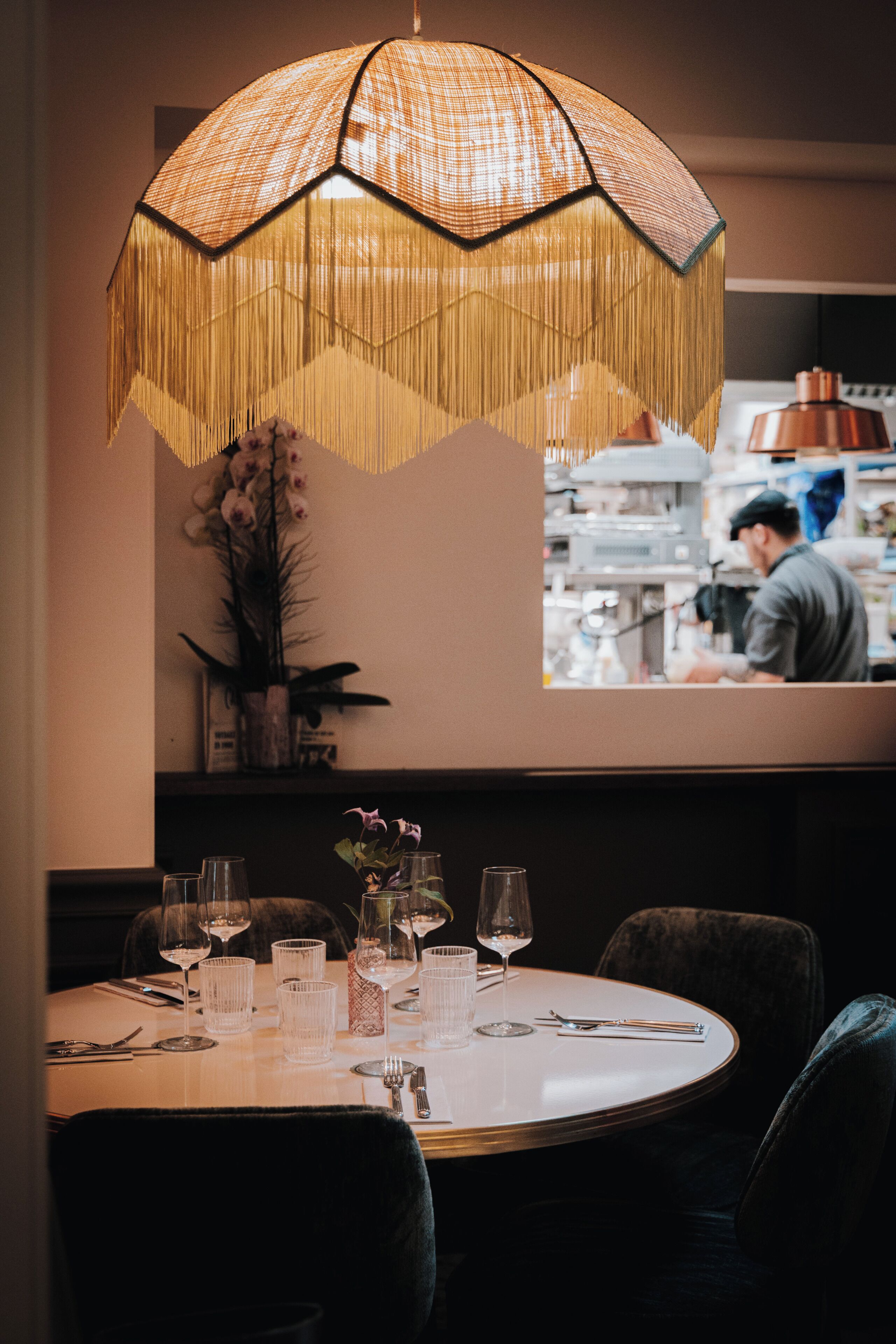 A dining area with a round table set for two, a hanging light fixture, and a view of a kitchen area.