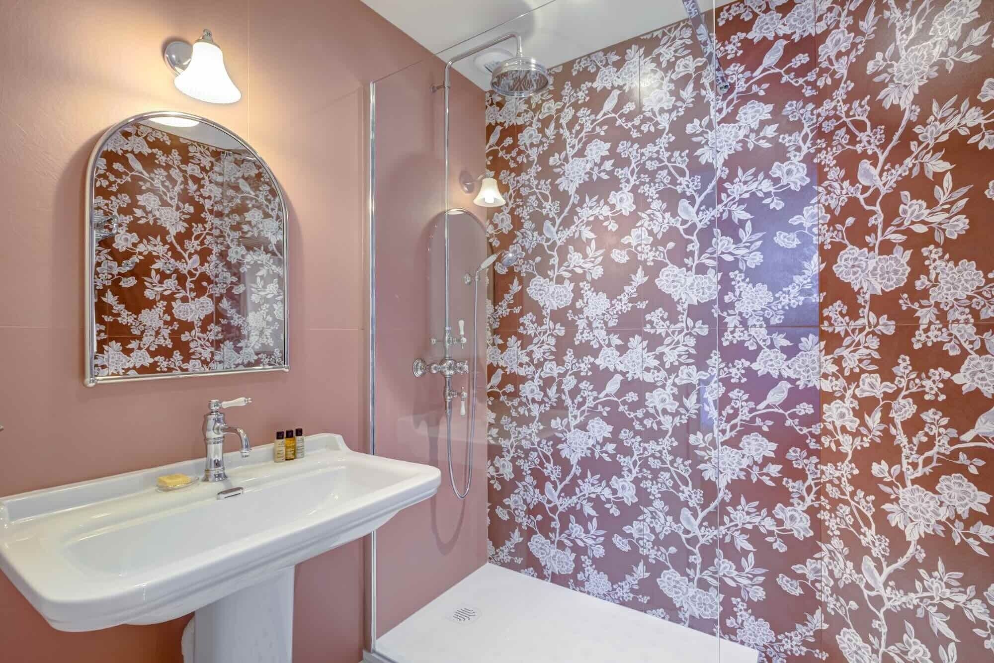 A bathroom with a sink, mirror, shower, and floral wallpaper.