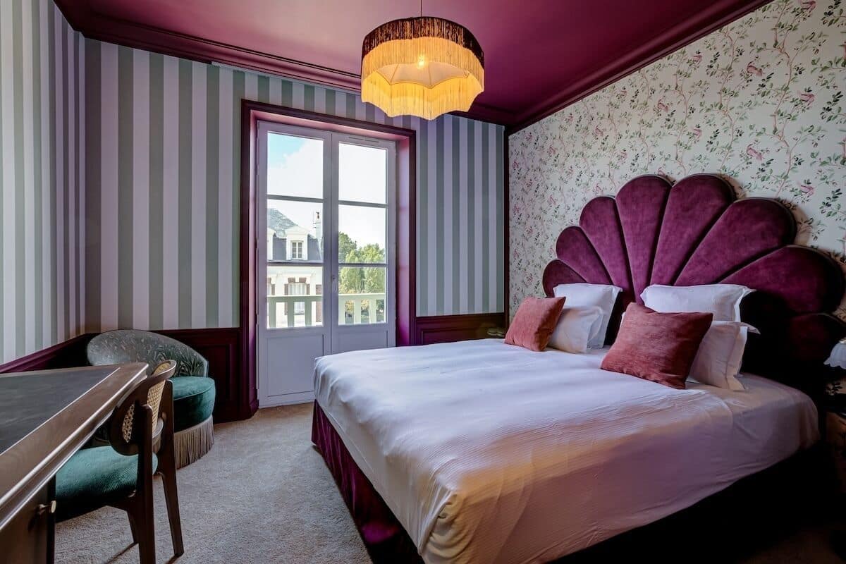 A hotel room with a bed, a desk, a chair, a window, and a chandelier.