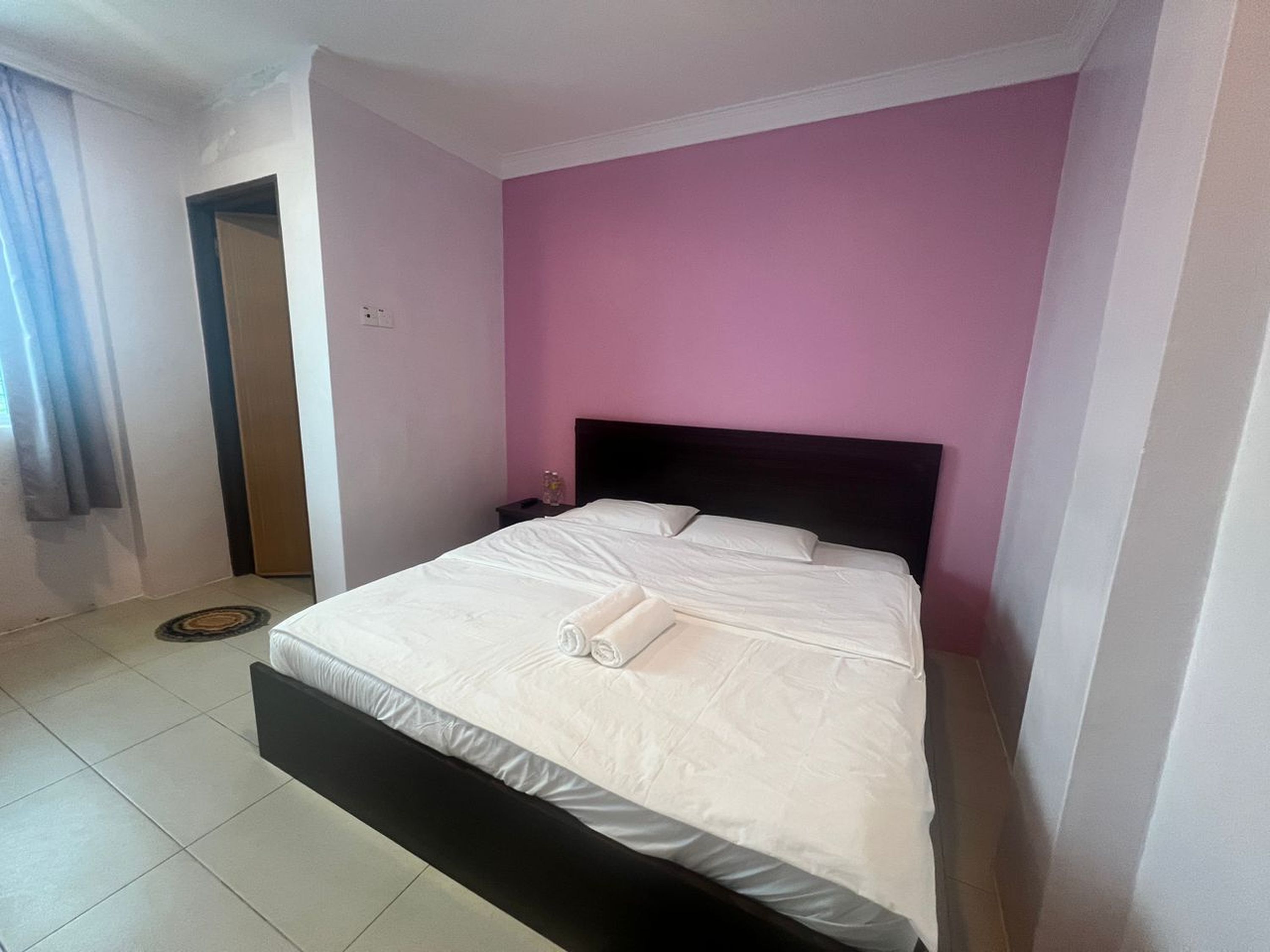 A neatly made bed with white linens and a rolled towel, a dark headboard, and a pink wall.