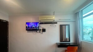 A room with a TV mounted on the wall, a small desk with a chair, a trash bin, and a window with a view.