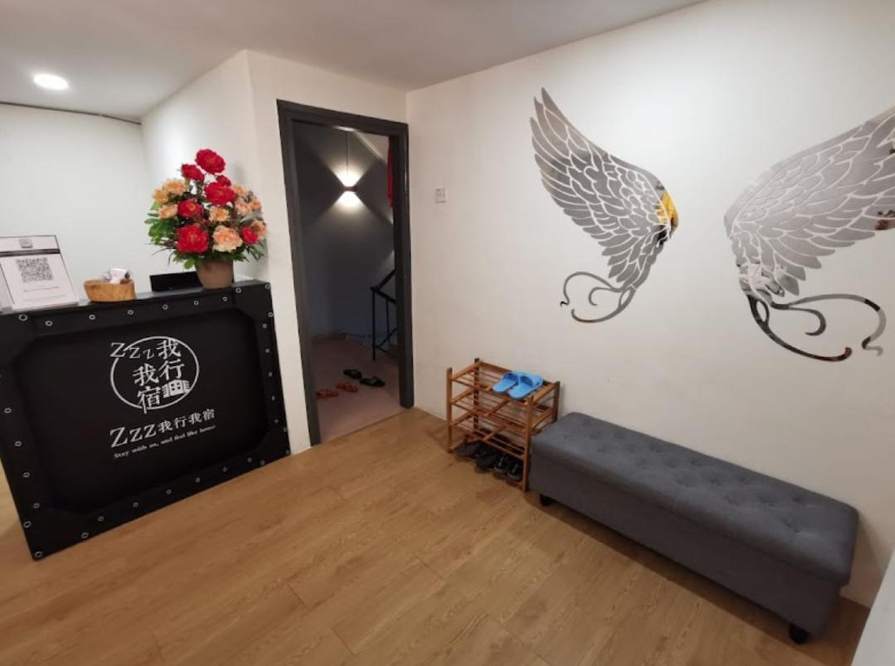 Een receptiezone met een houten vloer, een grijze bank, een bureau met zwart frame, een bloemstuk en wanddecoratie met vleugels.