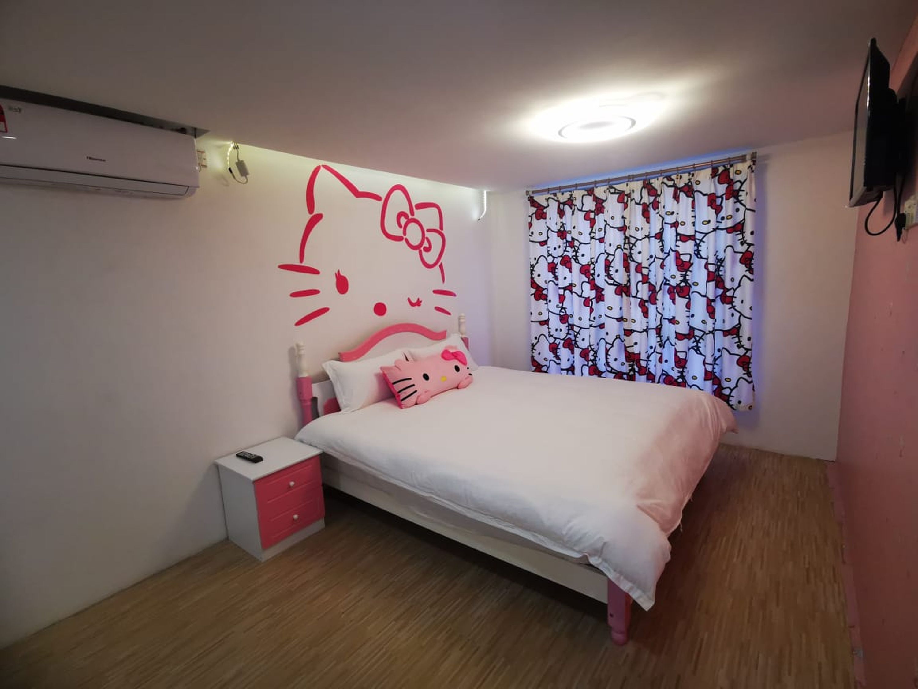 A bedroom with a bed, a nightstand, a small drawer unit, a window with curtains, and a wall-mounted TV.