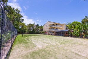 Cancha de tenis con red, rodeada por una cerca y abundante vegetación. Se ve un edificio de varios pisos con balcones al fondo.