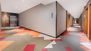 Un pasillo de hotel con alfombra de diseño geométrico, puertas de madera y un panel de control de ascensor en la pared.