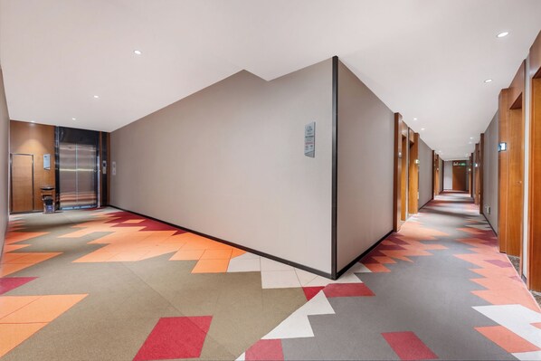 Ein Hotelkorridor mit einem geometrisch gemusterten Teppich, Holztüren und einem an der Wand montierten Bedienfeld für den Aufzug.