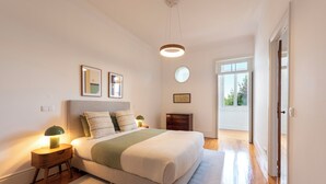 A bedroom with a bed, bedside tables, a nightstand, a wall-mounted lamp, framed pictures, and a view through an open door. - Bonfim 241 Family House (Porto)