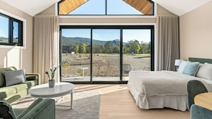 Ein modernes Hotelzimmer mit einem großen Bett, einer Couch, einem Esstisch und Blick auf eine gebirgige Landschaft durch raumhohe Fenster.