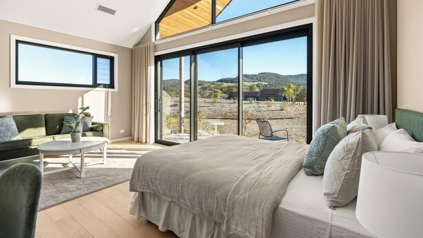 Ein modernes Schlafzimmer mit einem großen Bett, einem grünen Sofa, einem kleinen Tisch und Blick ins Freie durch große Fenster.