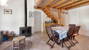 Una sala de estar acogedora con chimenea, vigas de madera en el techo, una mesa de comedor con sillas y una escalera que conduce a un nivel superior.