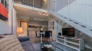 Ein modernes Wohnzimmer mit einer Couch, einem Couchtisch und einer TV-Konsole. Eine Treppe führt zu einer oberen Ebene mit einer Küchenzeile.