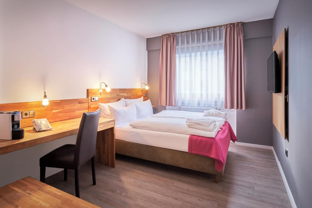 A hotel room with a wooden desk, a bed with a pink blanket, a TV mounted on the wall, and a window with curtains.