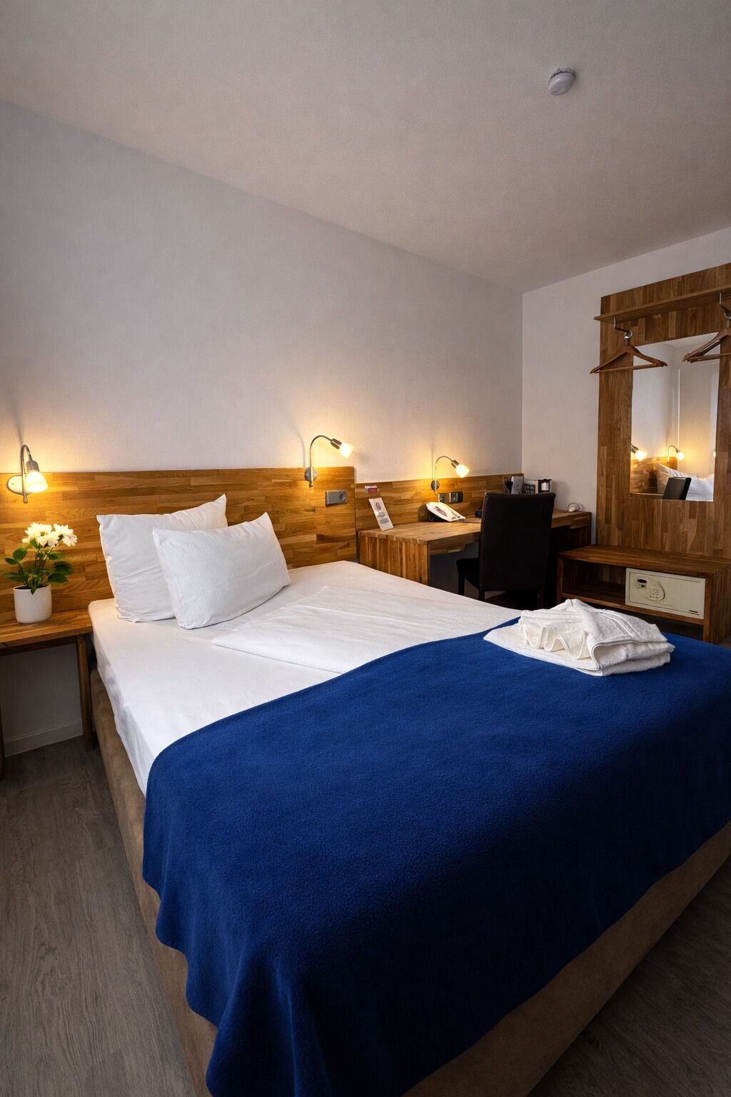 Ein Hotelzimmer mit einem hölzernen Kopfteil, einem Bett mit weißen Leinen, einer blauen Tagesdecke, einem Schreibtisch mit Telefon und einem Spiegel mit Holzrahmen.