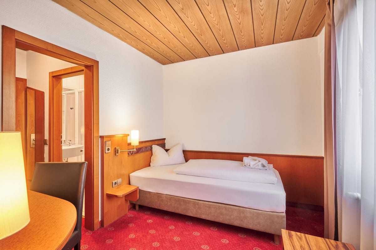 A hotel room with a wooden ceiling, a bed with white linens, a red carpet, a wooden nightstand, and a table with a lamp.