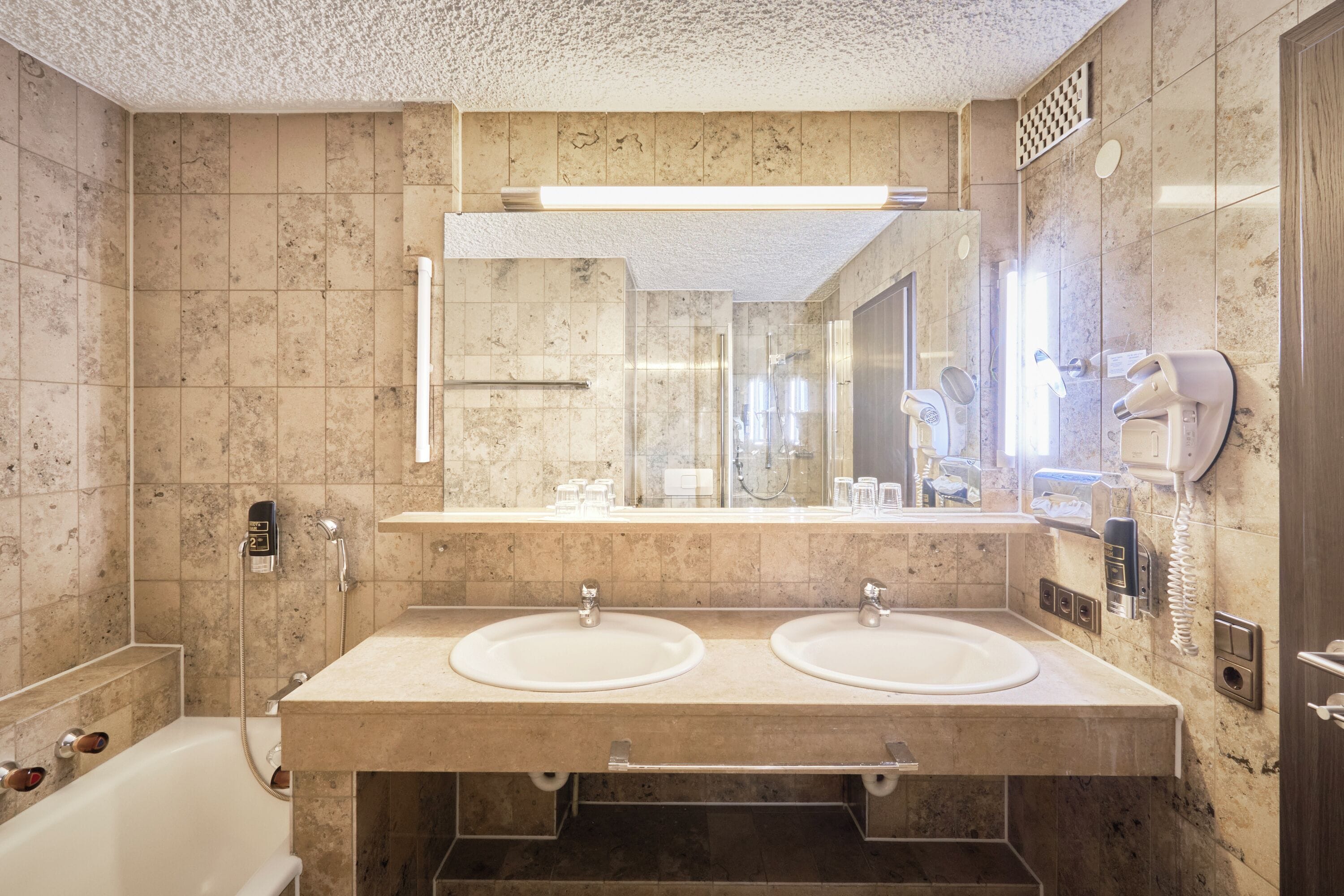 A bathroom with two sinks, a large mirror, a shower, and a hairdryer on the wall.