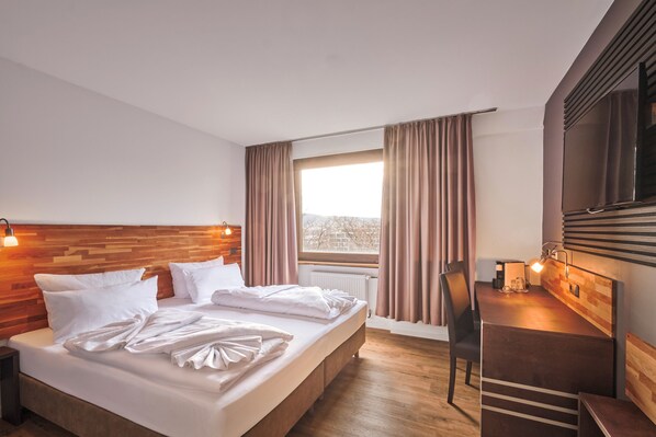 A hotel room with a wooden headboard, a large bed with white linens, a desk with a chair, and a large window with curtains. - Luna Inn (Stuttgart)