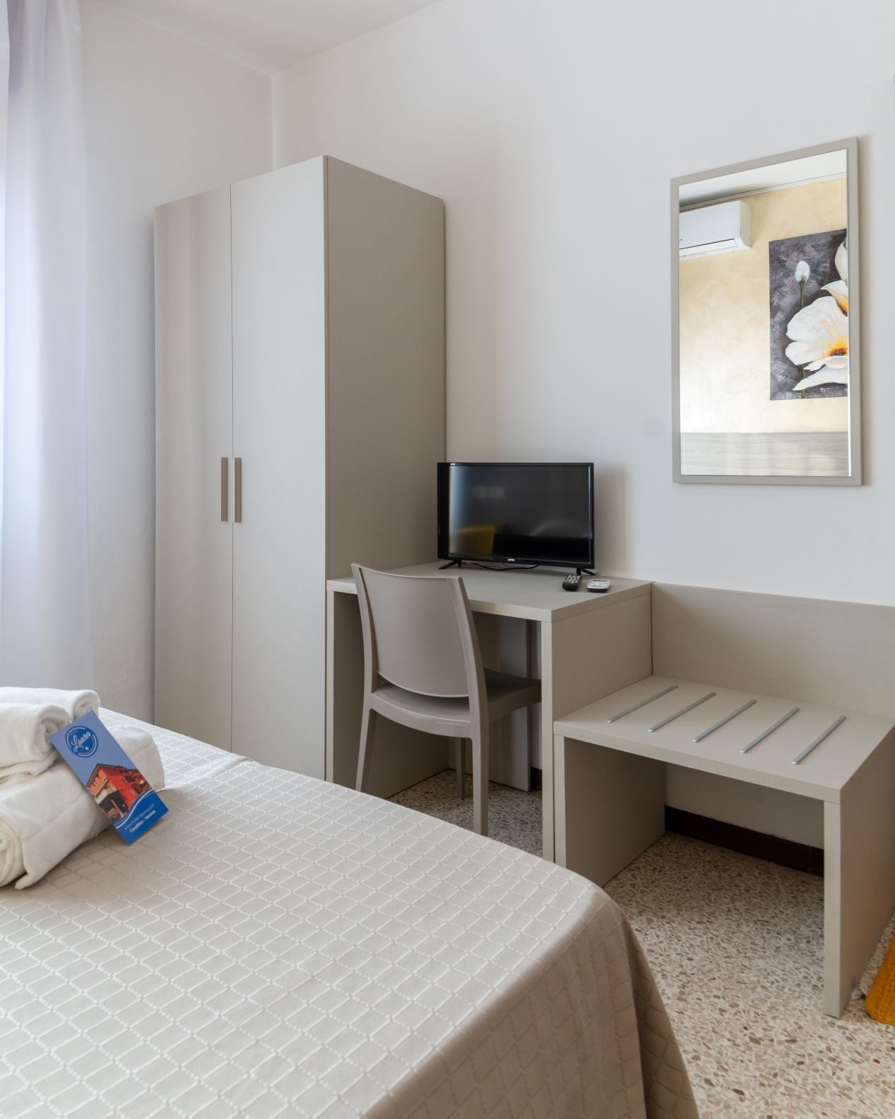 A hotel room with a bed, a desk with a computer, a chair, a wardrobe, and a painting on the wall.