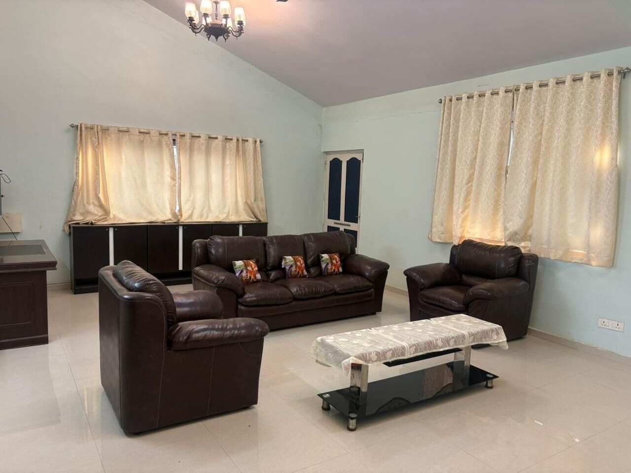 A living room with a brown leather sofa set, a coffee table, and a TV. There are curtains on the windows, and a chandelier hangs from the ceiling.