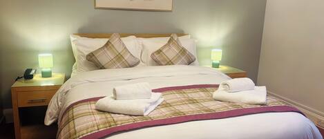 A neatly made bed with a plaid bedspread, two pillows, and two folded towels on the bed. There are two bedside tables with lamps, a wall-mounted artwork, and a telephone on one of the tables.