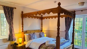 A four-poster bed with a wooden frame, a bedside table with a lamp, and a view of greenery through a window.